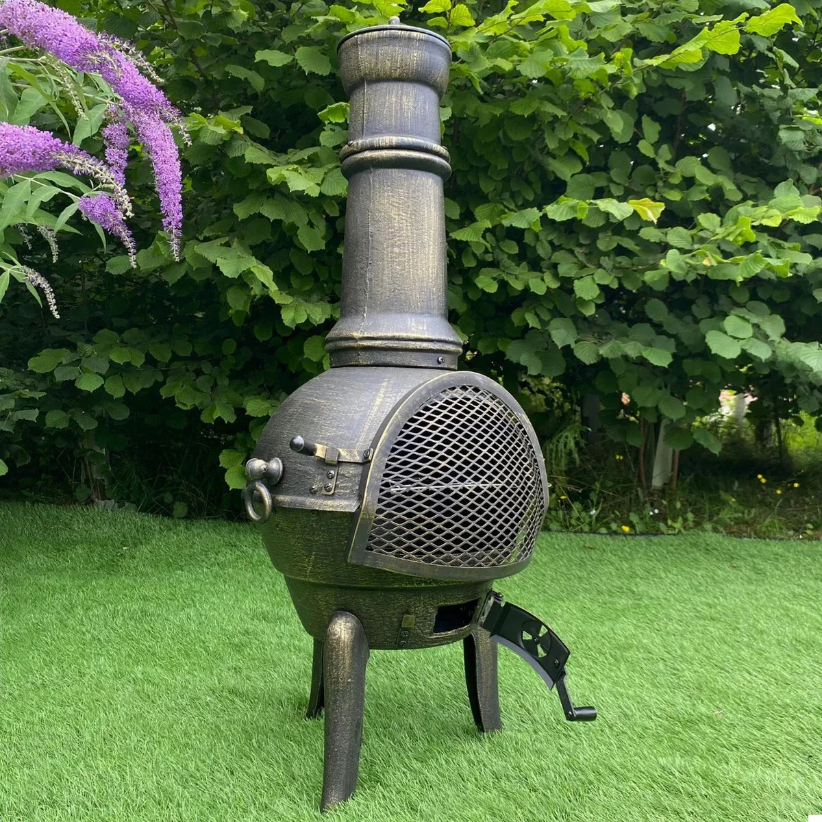 Cast iron chimenea with a textured black finish, featuring a tall chimney, mesh door, and sturdy legs, set against lush greenery