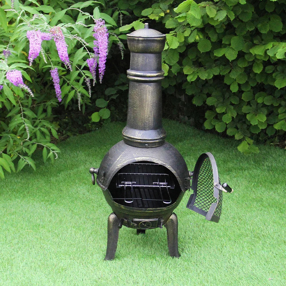 Cast iron chimenea with a tall flue, textured bronze finish, and open door revealing a grill, set against lush greenery and purple flowers