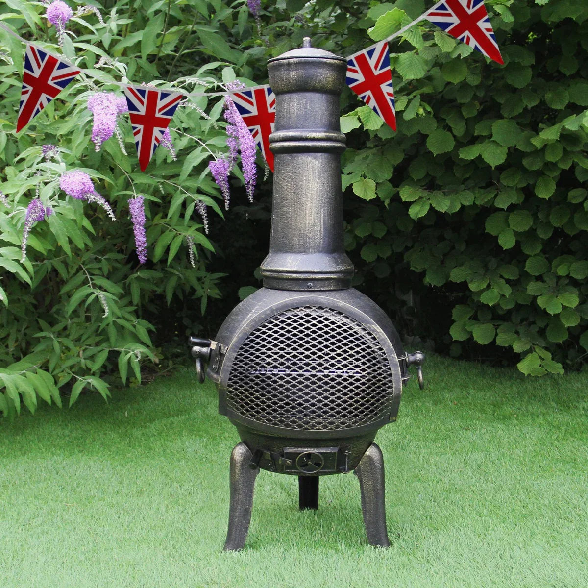 Cast iron chimenea with a tall chimney, mesh front, and Union Jack bunting, set against lush green foliage and grass