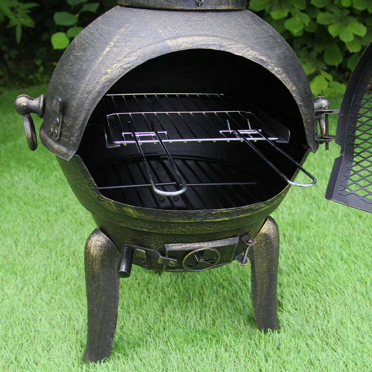 Cast iron chimenea with an open door revealing a spacious cooking grate, featuring a rustic bronze finish and sturdy legs
