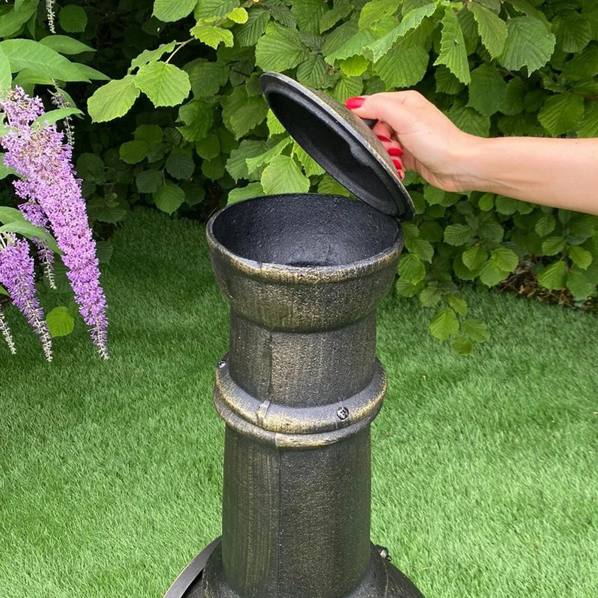 Cast iron chimenea with a hand lifting the lid, surrounded by lush greenery and purple flowers, showcasing its rustic texture and design