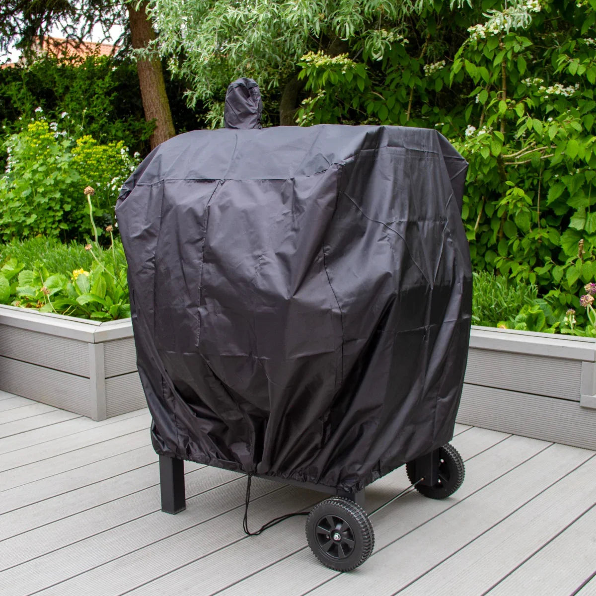 XL BBQ smoker grill covered with a black protective cover, featuring wheels for mobility, set on a wooden deck surrounded by greenery
