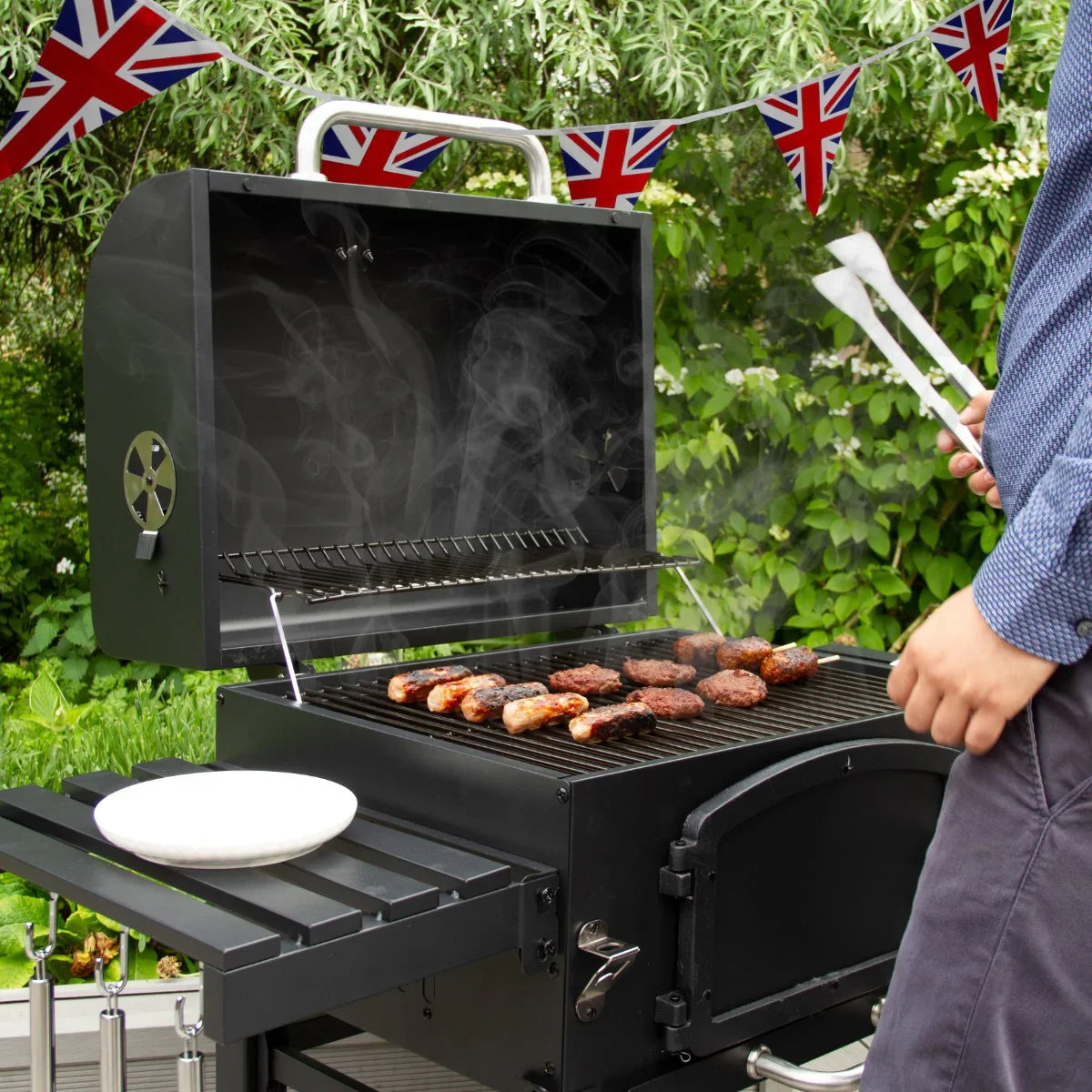 XL BBQ smoker grill with smoky sausages and burgers, set outdoors with Union Jack bunting in a lush garden backdrop