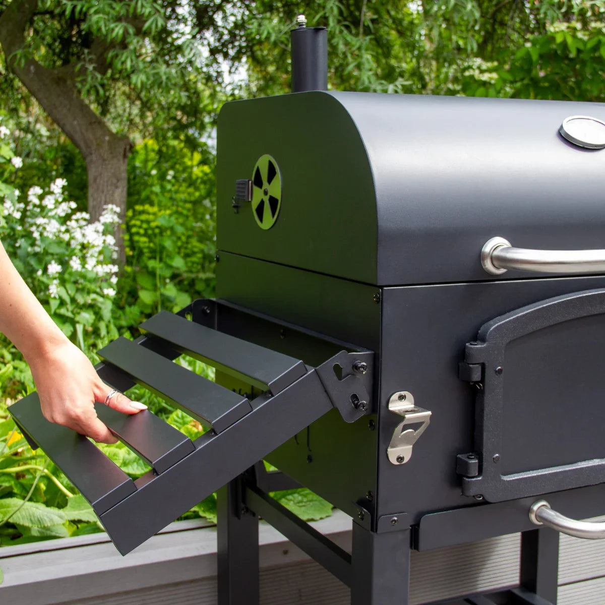 XL BBQ smoker grill in sleek black with a side shelf being pulled out, surrounded by lush greenery and white flowers
