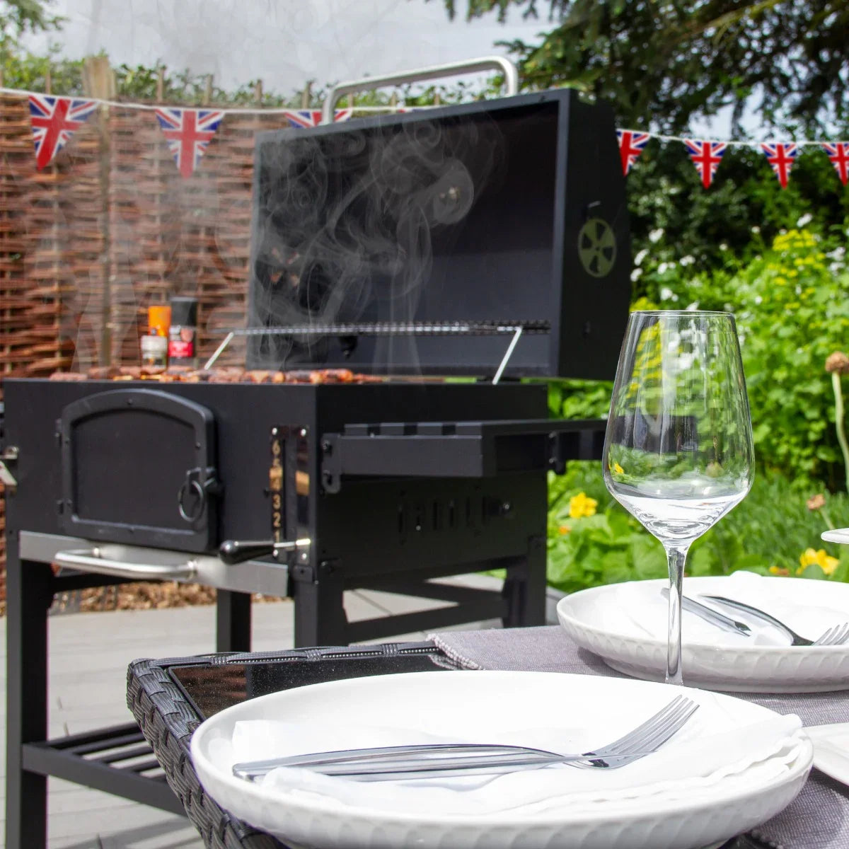 XXL BBQ smoker grill with smoke rising, Union Jack bunting in background, set on patio with plates and glass ready for outdoor cooking
