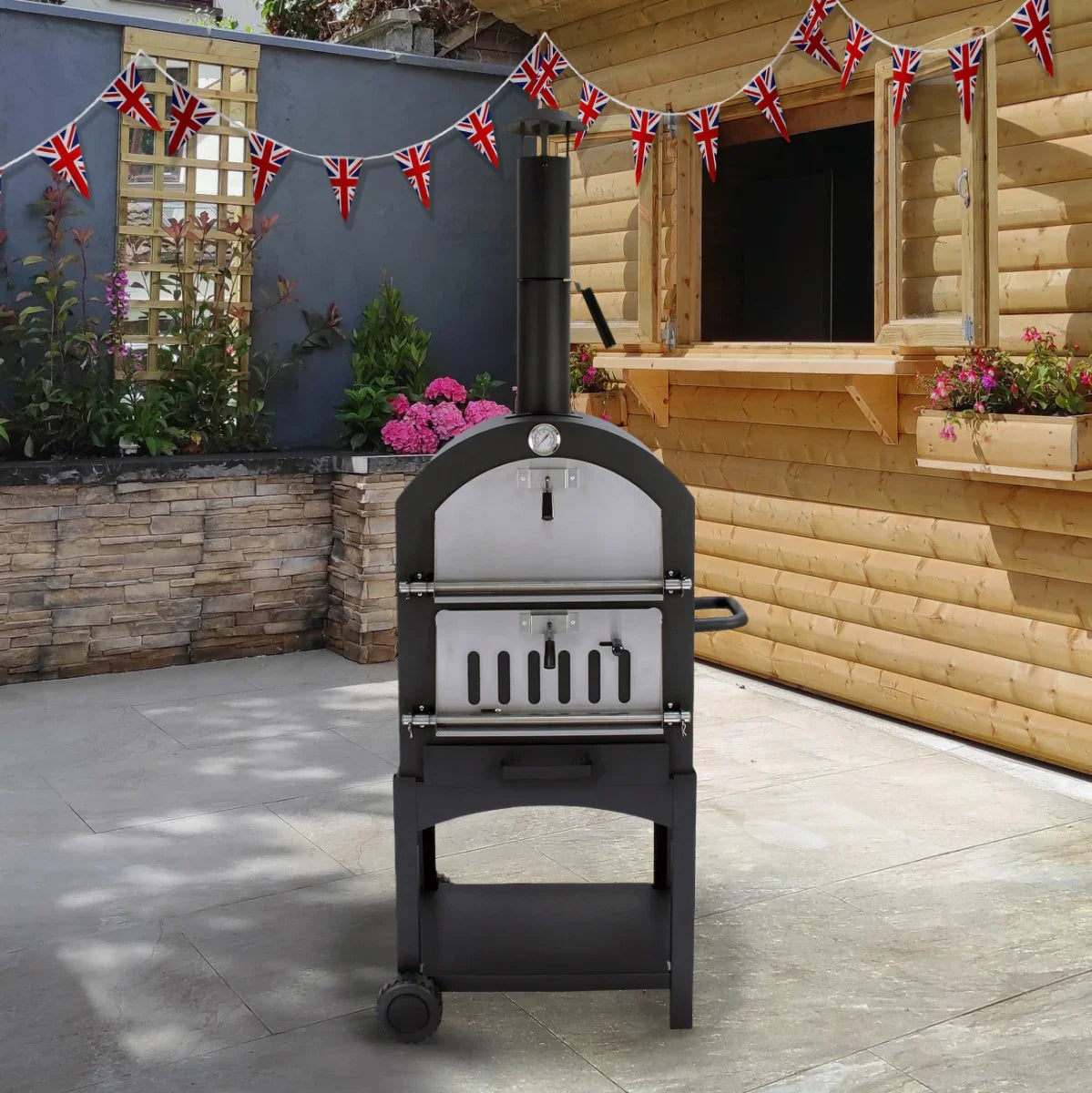 Outdoor pizza oven with a sleek black finish, stainless steel details, and a rain cover, set against a rustic wooden backdrop with Union Jack bunting