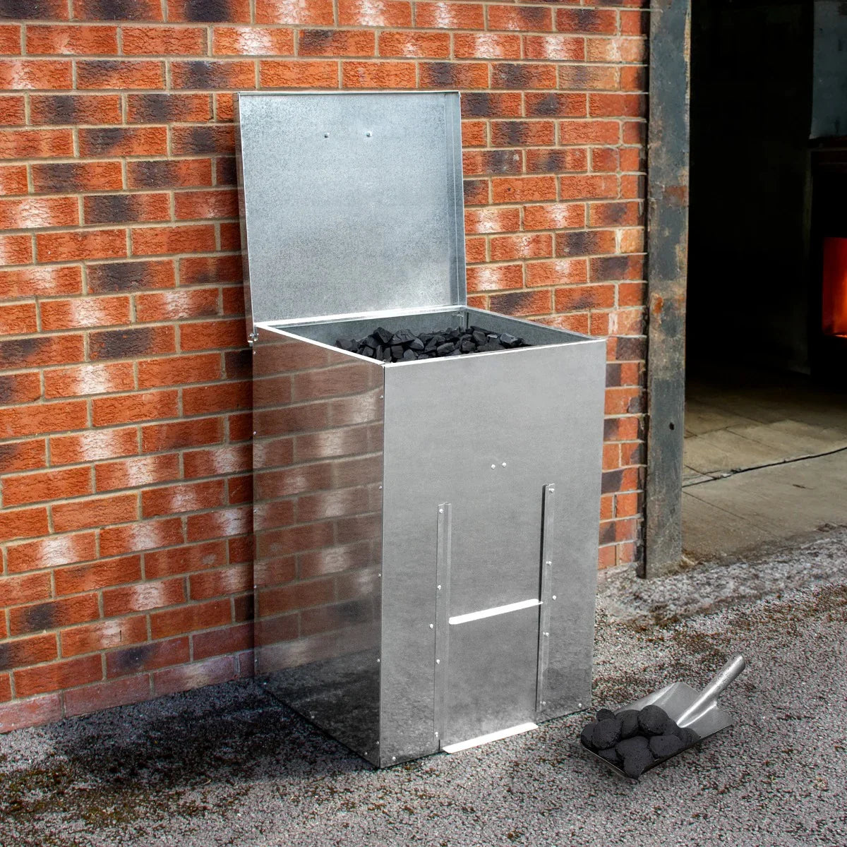 Galvanised coal bunker with a shiny metallic finish, holding black coal, set against a brick wall and gravel ground