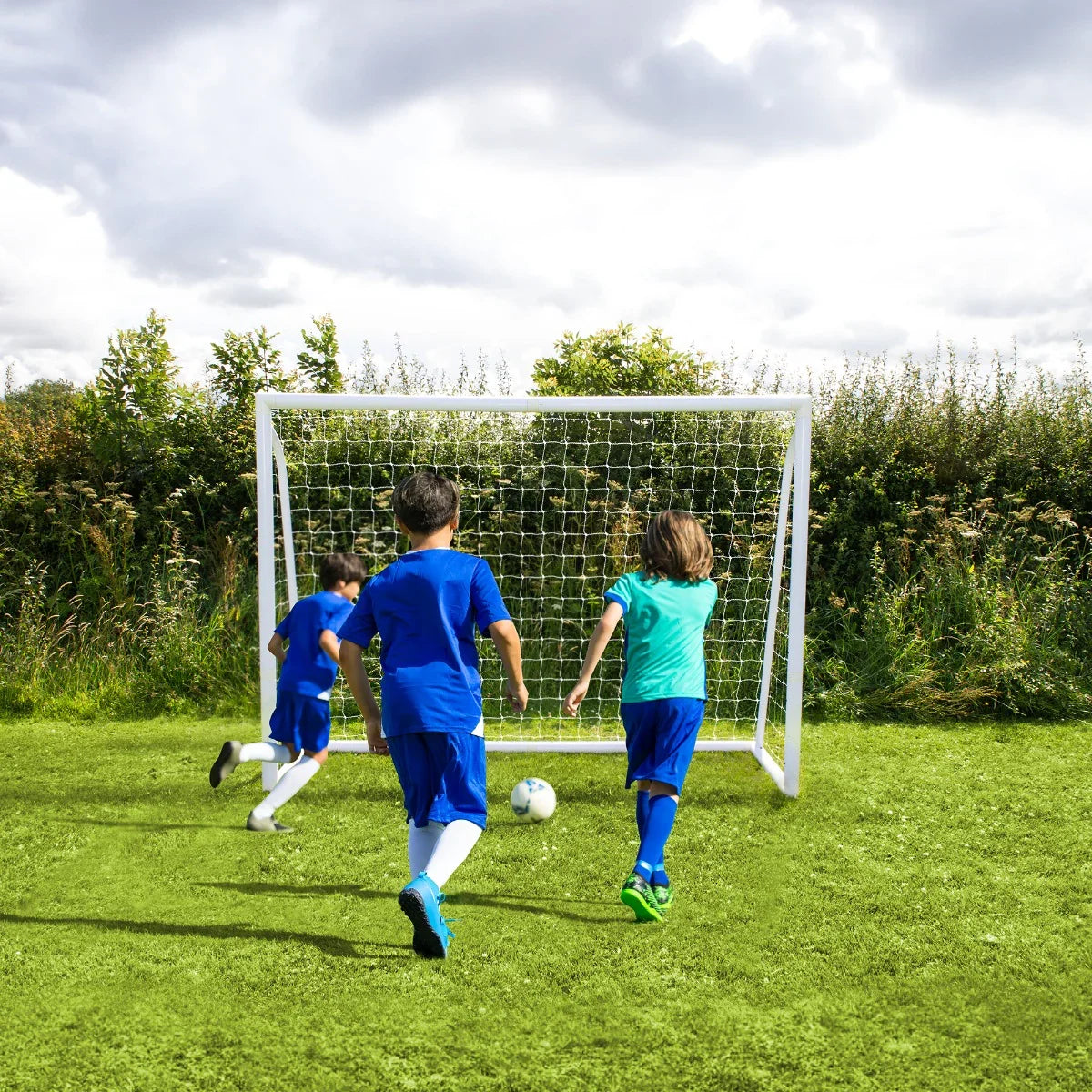Children in blue soccer uniforms play near an 8 x 6ft football goal on a grassy field, with a cloudy sky in the background