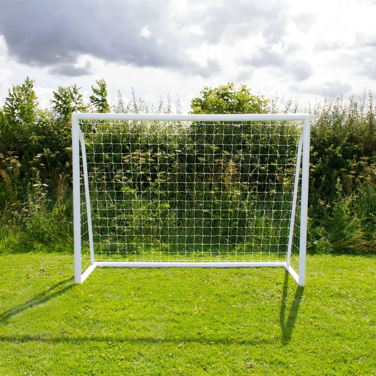 8 x 6ft white football goal with a sturdy net, set on green grass, surrounded by lush greenery and a cloudy sky
