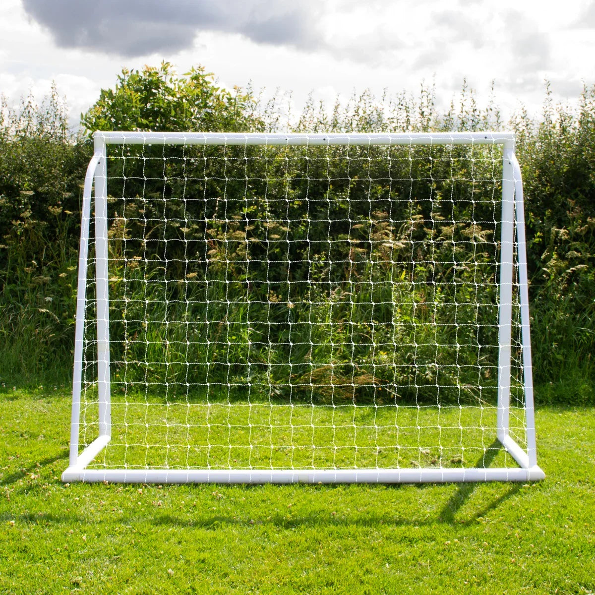 8 x 6ft football goal with white frame and netting, set on green grass, surrounded by lush greenery under a cloudy sky