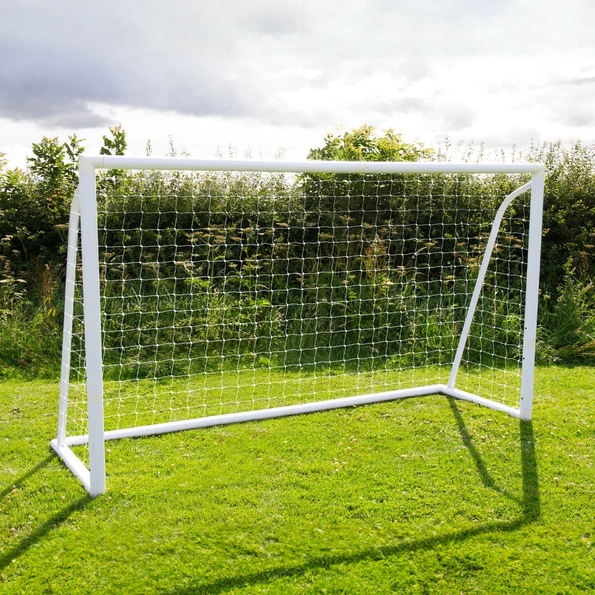 12 x 6ft white football goal with netting, set on green grass, surrounded by lush greenery under a cloudy sky