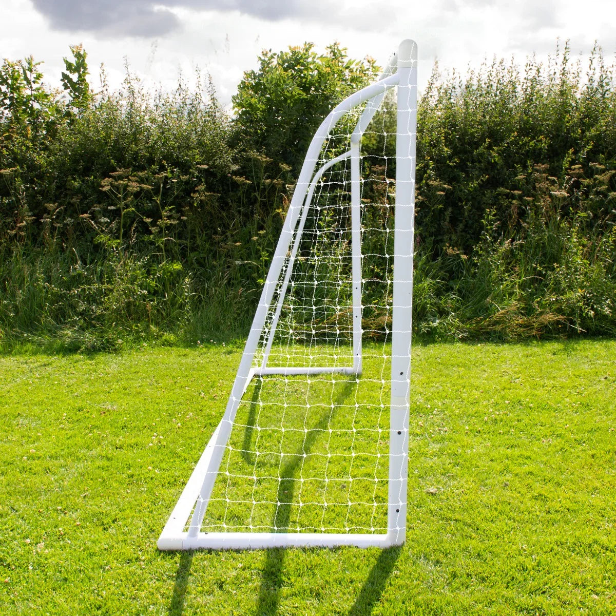 12 x 6ft white football goal with netting, angled view on green grass, surrounded by tall grass and trees in background