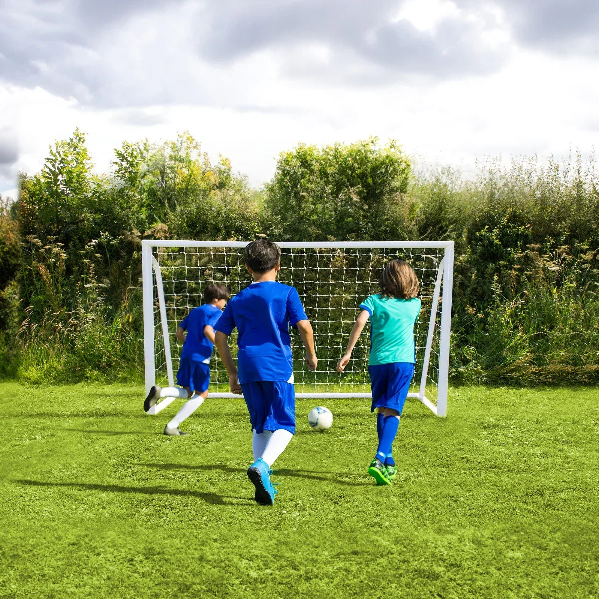 Three children in blue jerseys play soccer, running towards a white 6 x 4ft football goal on a grassy field under a cloudy sky