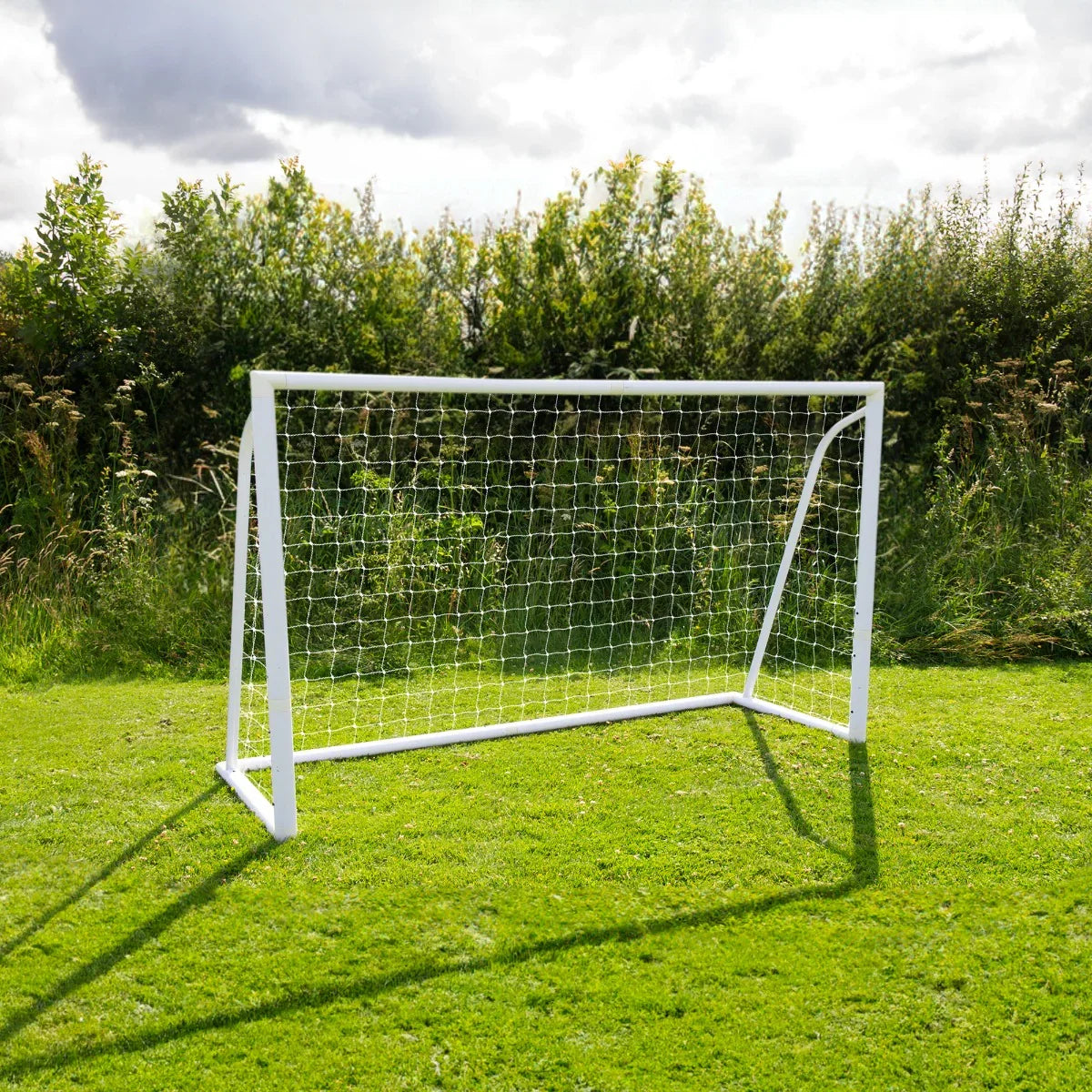 6 x 4ft white football goal with netting, set on green grass, surrounded by lush greenery under a cloudy sky