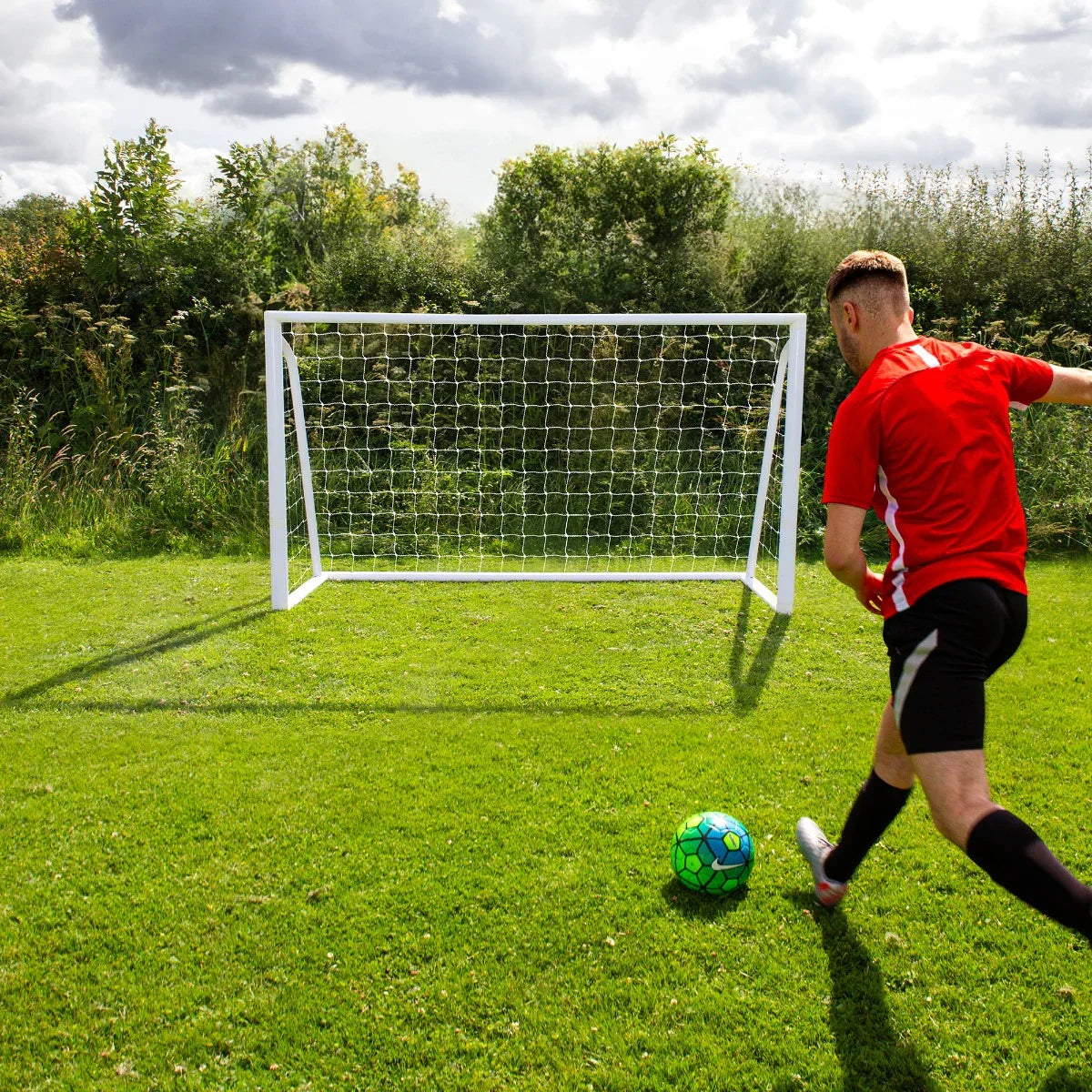 6 x 4ft football goal with white frame and net, set on green grass, as a player in red kicks a colorful soccer ball towards it