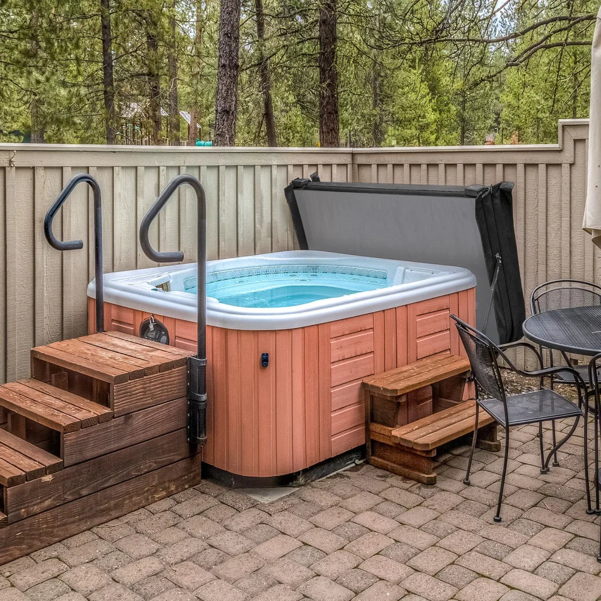 Black 2m hot tub spa cover partially opened, showcasing the spa's water, surrounded by wooden steps and a stone patio