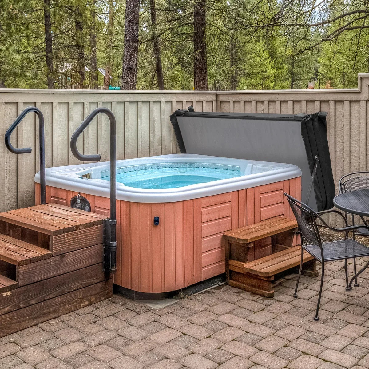 Black 2.1m hot tub spa cover resting on a wooden hot tub, surrounded by a stone patio and trees, with steps leading up to it