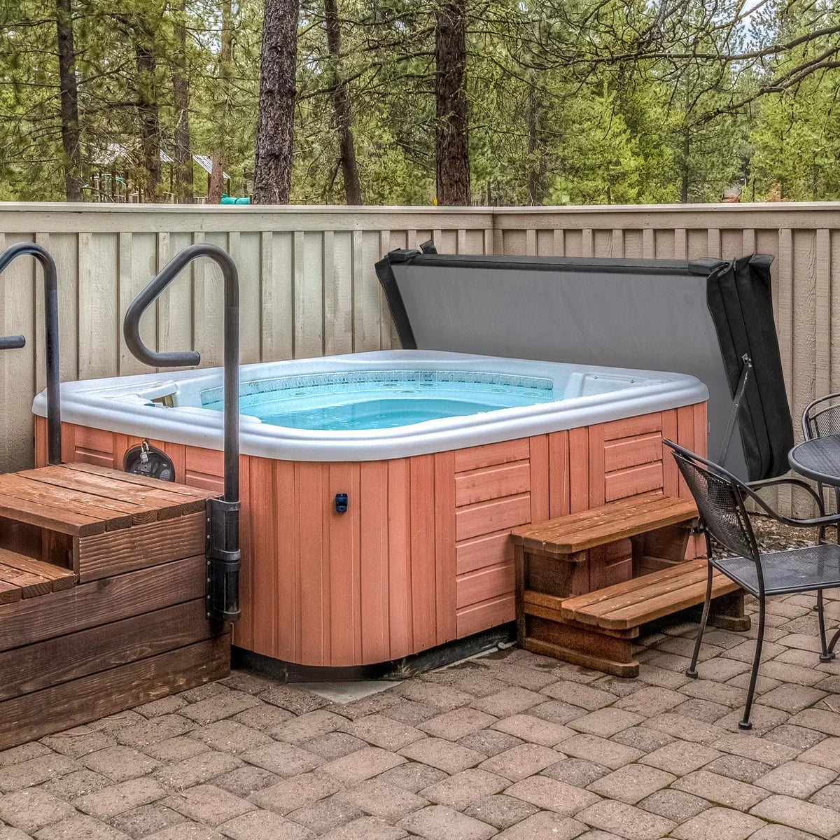 Black 2.2m hot tub spa cover resting on a wooden hot tub, surrounded by a stone patio and lush green trees in the background