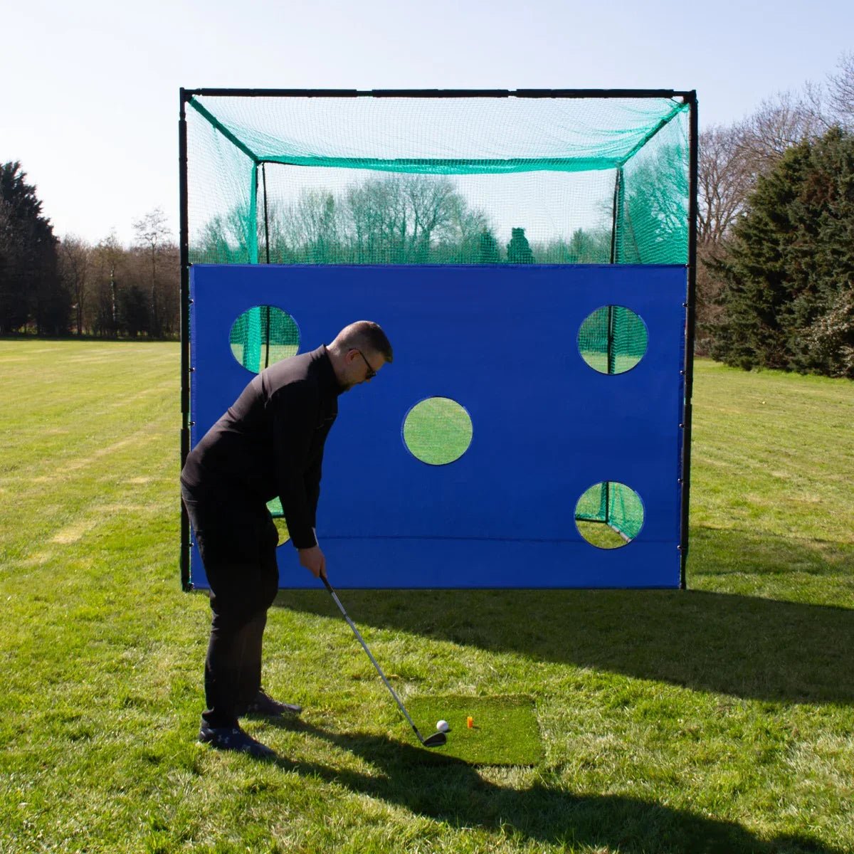 Golf practice cage with a blue target sheet featuring circular holes, set on a grassy field with a golfer preparing to swing