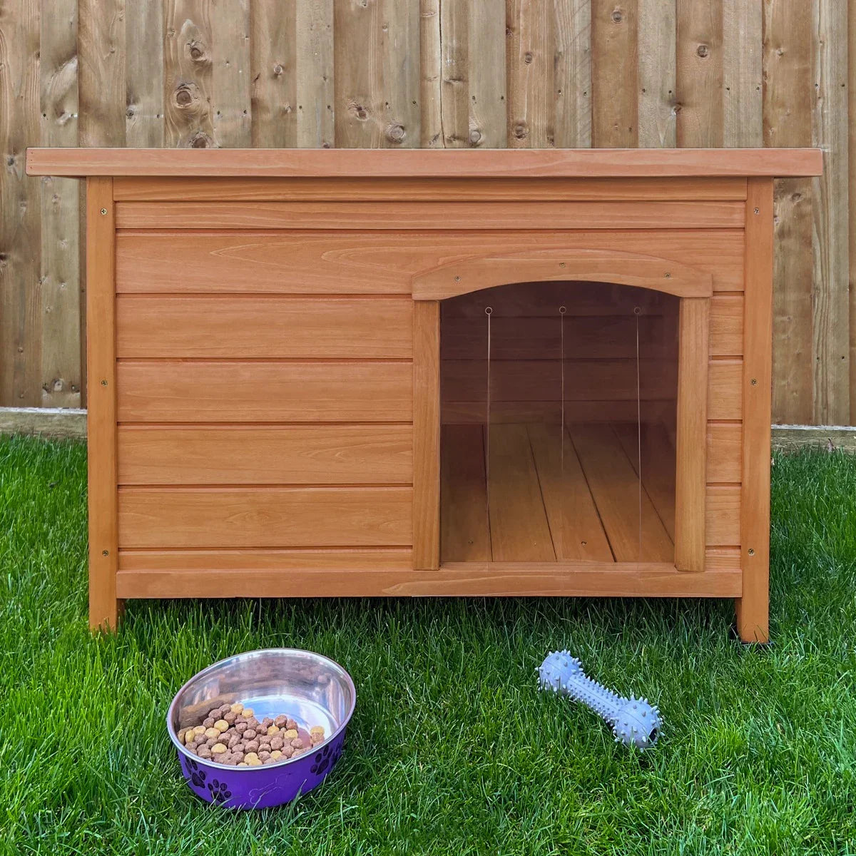 Small wooden dog kennel with a slanted roof, featuring a cozy entrance, placed on green grass beside a bowl of dog food and a toy