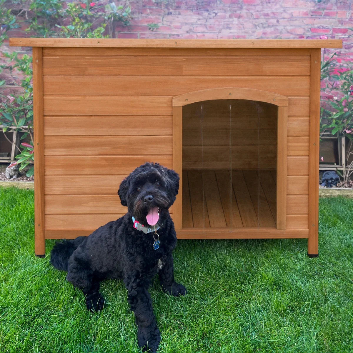 Large wooden dog kennel with a slanted roof, featuring a spacious entrance, placed on green grass with a black dog sitting nearby