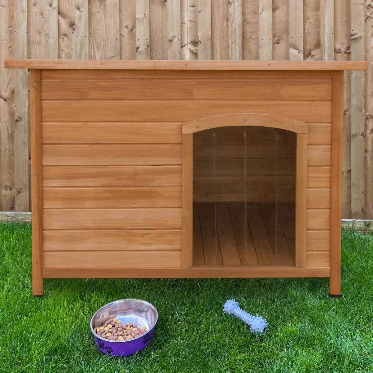 Large wooden dog kennel with a sloped roof, natural wood finish, and an open front, set on green grass beside a food bowl and chew toy