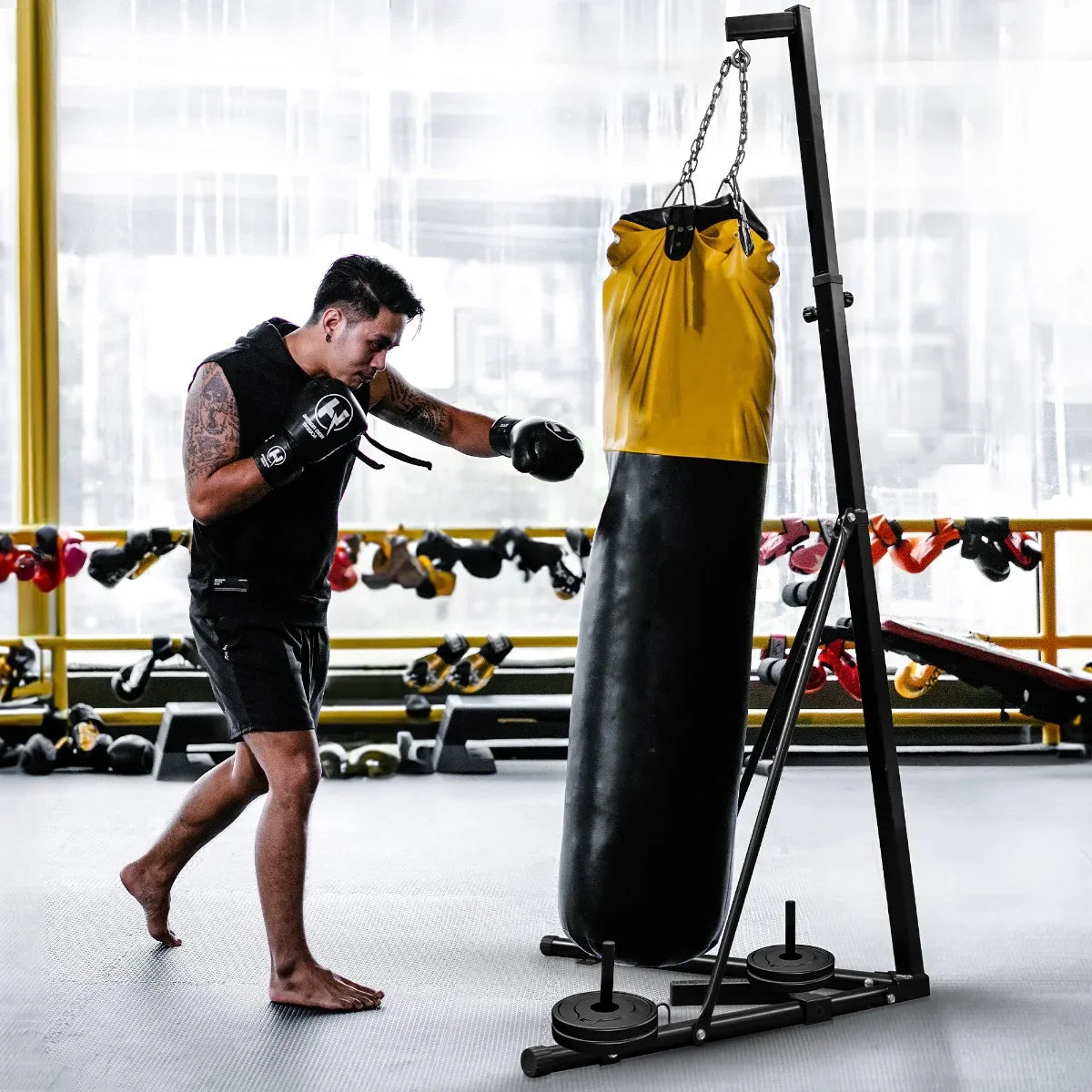 Folding punch bag stand with a black and yellow heavy bag, sturdy frame, and weights, set in a gym with colorful equipment in the background