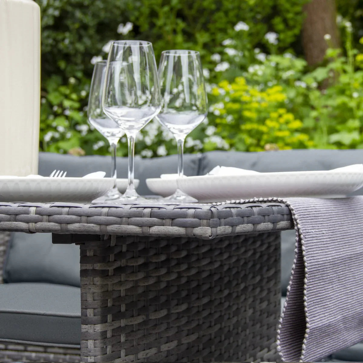 Grey rattan corner set featuring a textured finish, accompanied by elegant glassware and tableware, set against a lush garden backdrop