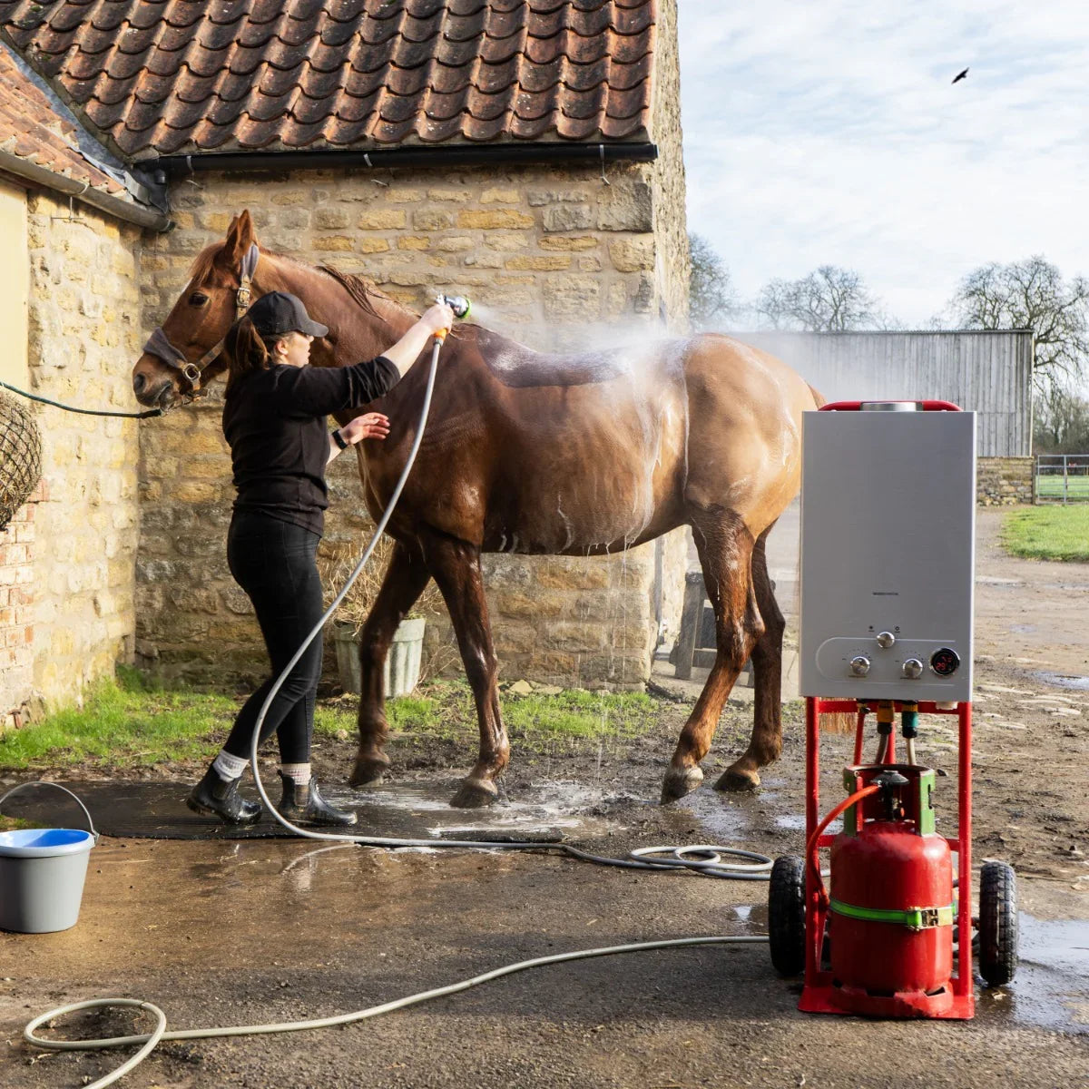 Portable horse shower in red, 18L capacity, with a woman washing a brown horse outdoors near a stone barn, water spraying