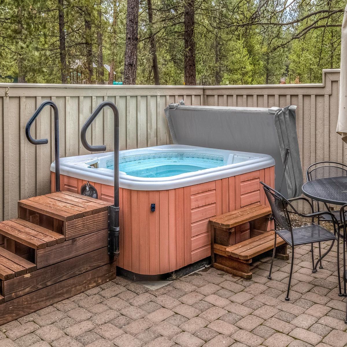 Grey 2m hot tub spa cover resting on a wooden hot tub, surrounded by a stone patio and wooden steps in a forested setting
