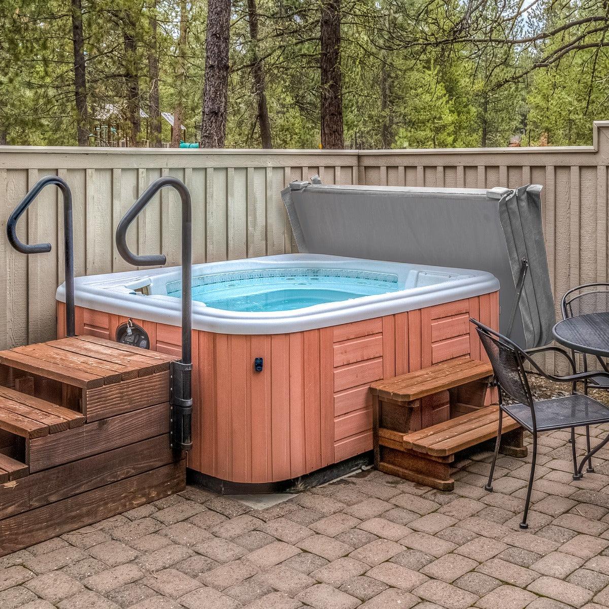 Grey 2.1m hot tub spa cover resting on a wooden hot tub, surrounded by a stone patio and wooden steps, with trees in the background