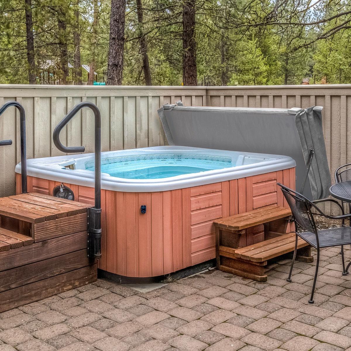 Grey 2.2m hot tub spa cover resting on a wooden hot tub, surrounded by a stone patio and outdoor seating area in a forested setting
