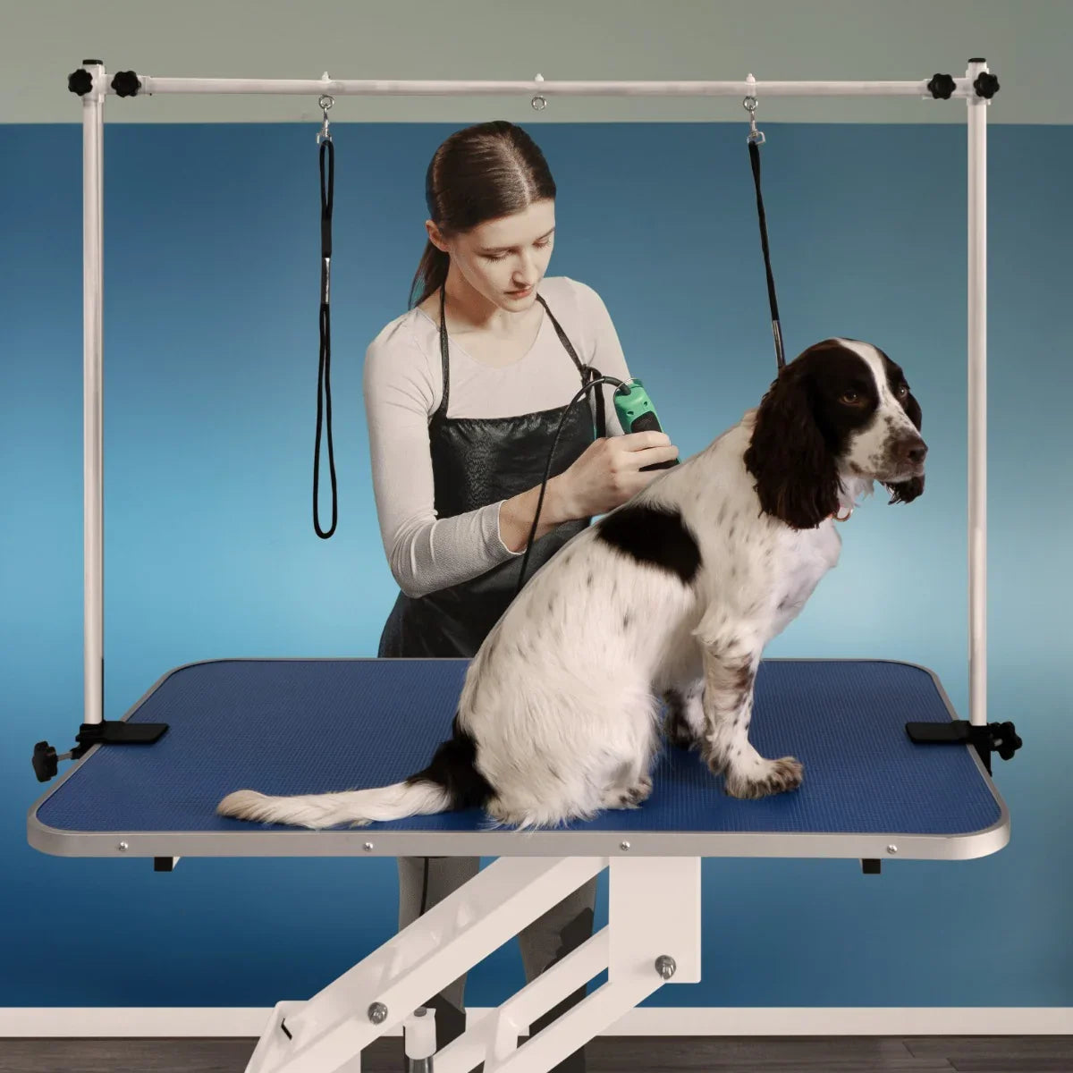 White hydraulic grooming table with a blue textured top, featuring a dog being groomed by a stylist using clippers