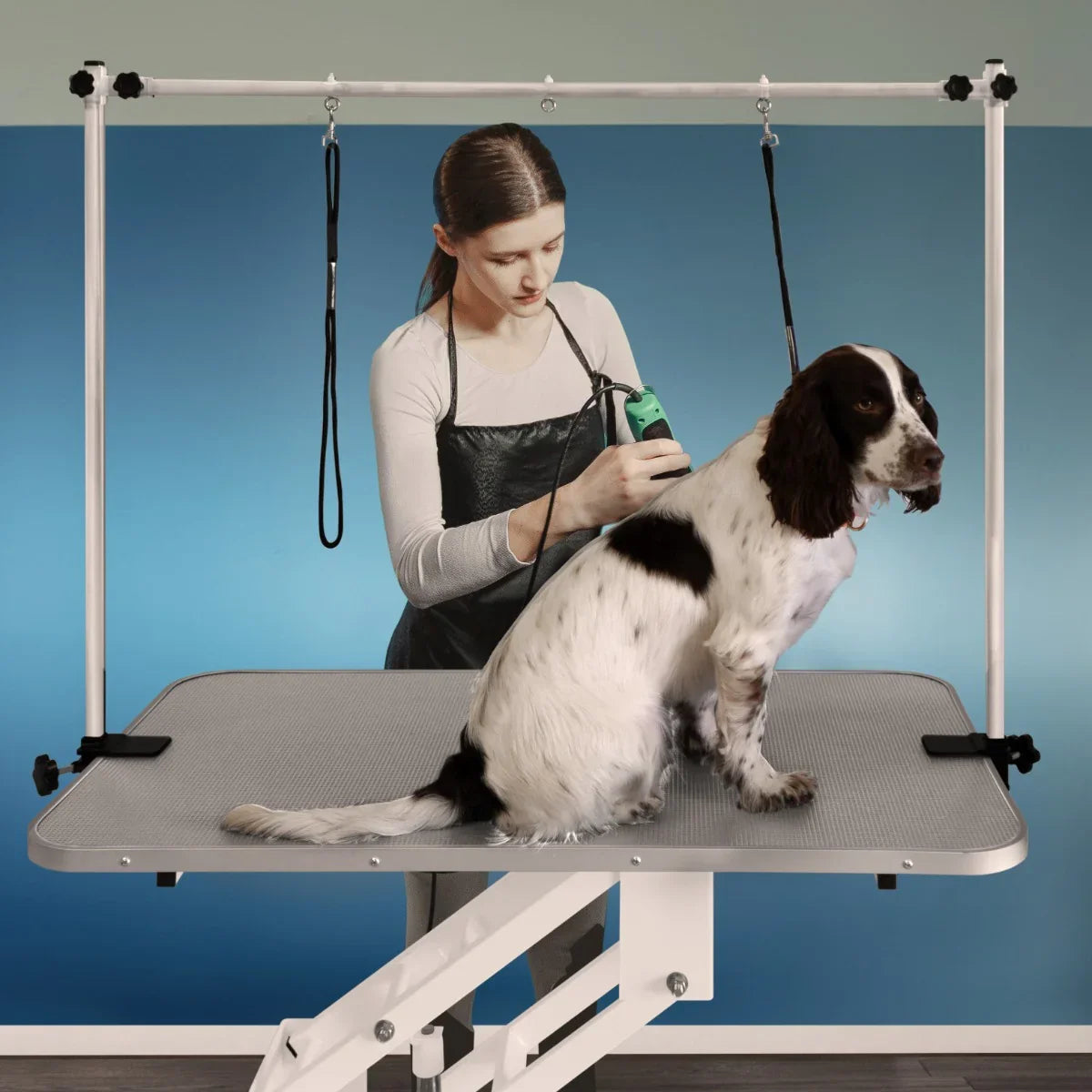 White hydraulic grooming table with a grey textured top, adjustable height, and a groomer using clippers on a dog