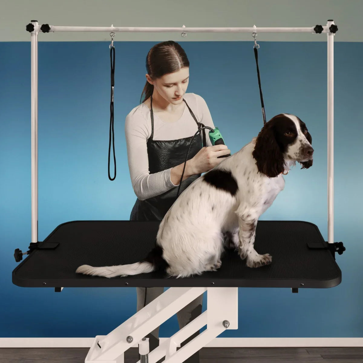 White hydraulic grooming table with a black textured top, featuring a dog being groomed by a stylist in a modern setting