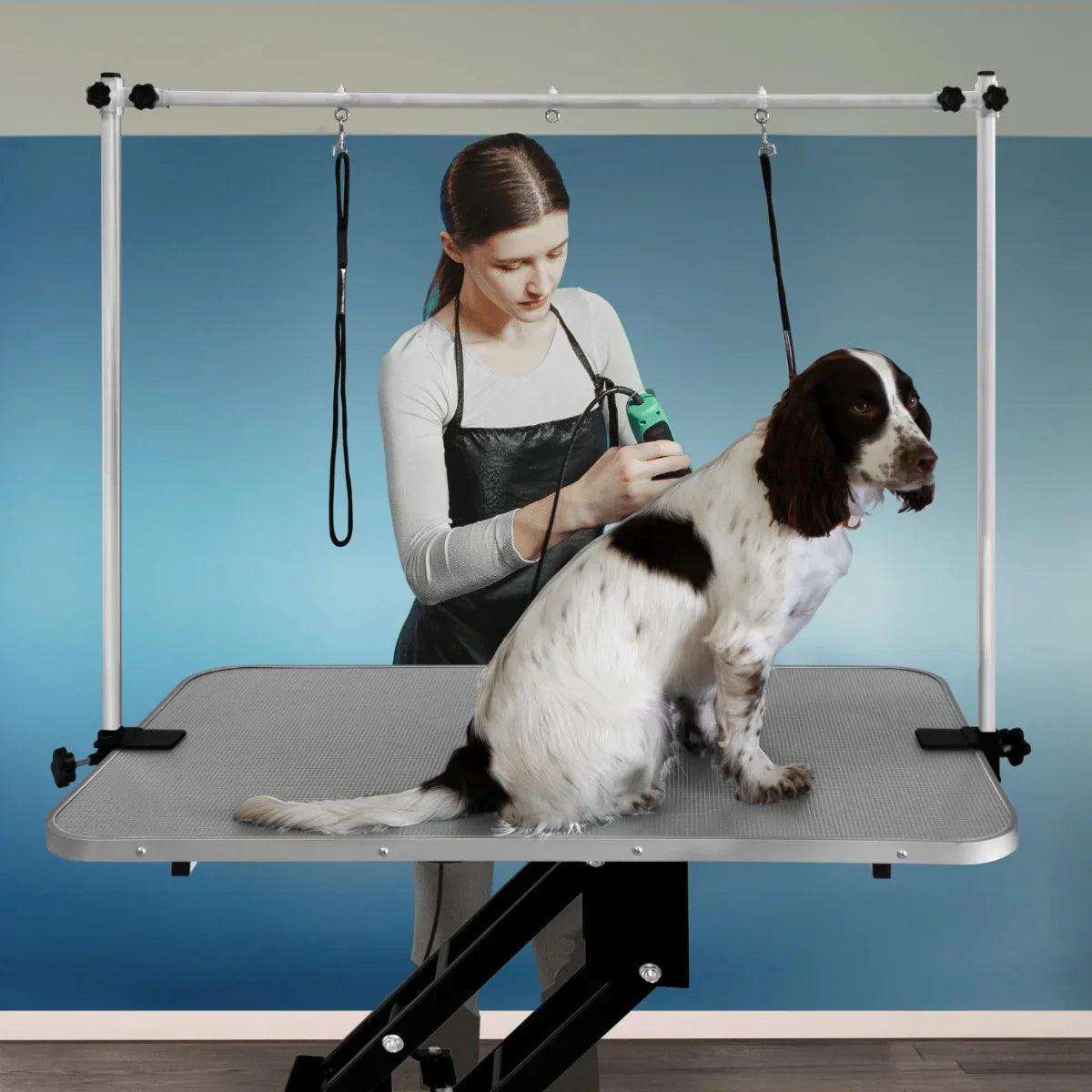 Black hydraulic grooming table with a grey textured top, adjustable height, and a dog being groomed by a stylist in the background