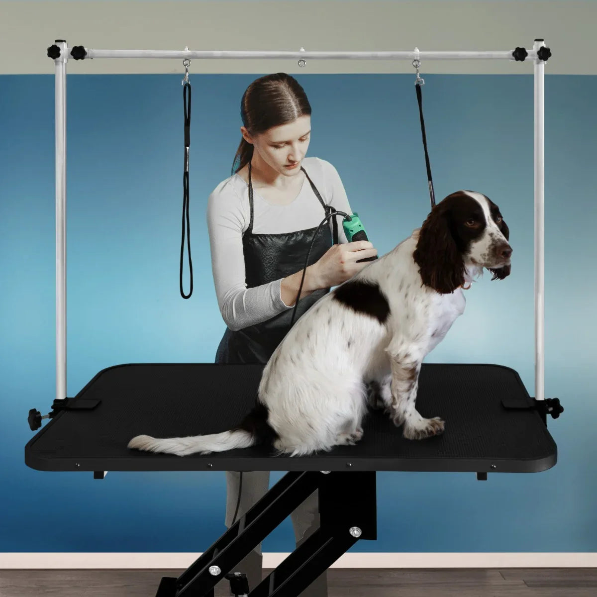 Black hydraulic grooming table with a textured surface, adjustable height, and a dog being groomed by a stylist in the background