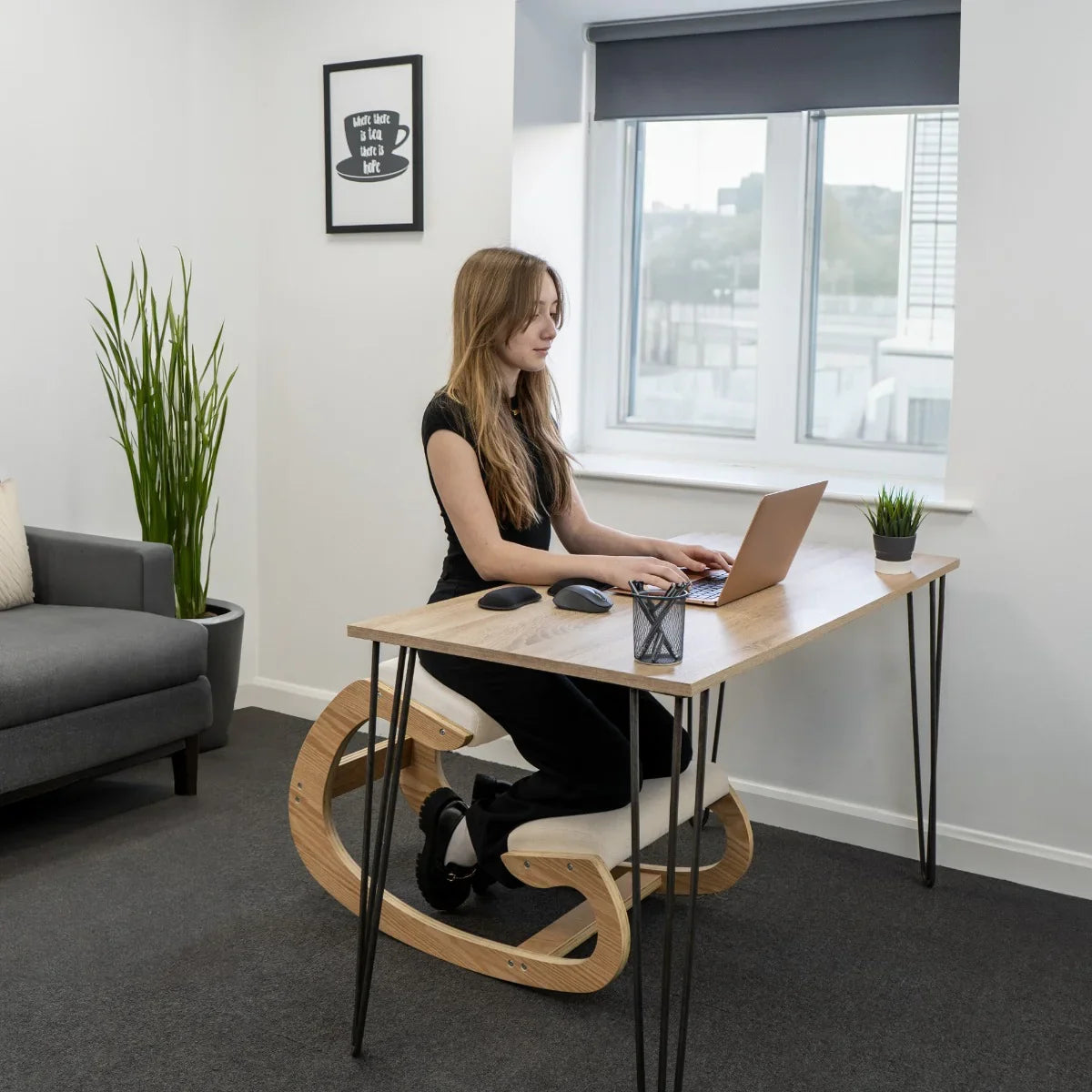 Ergonomic kneeling chair with a curved wooden base, supporting a user typing on a laptop at a modern desk in a bright office setting