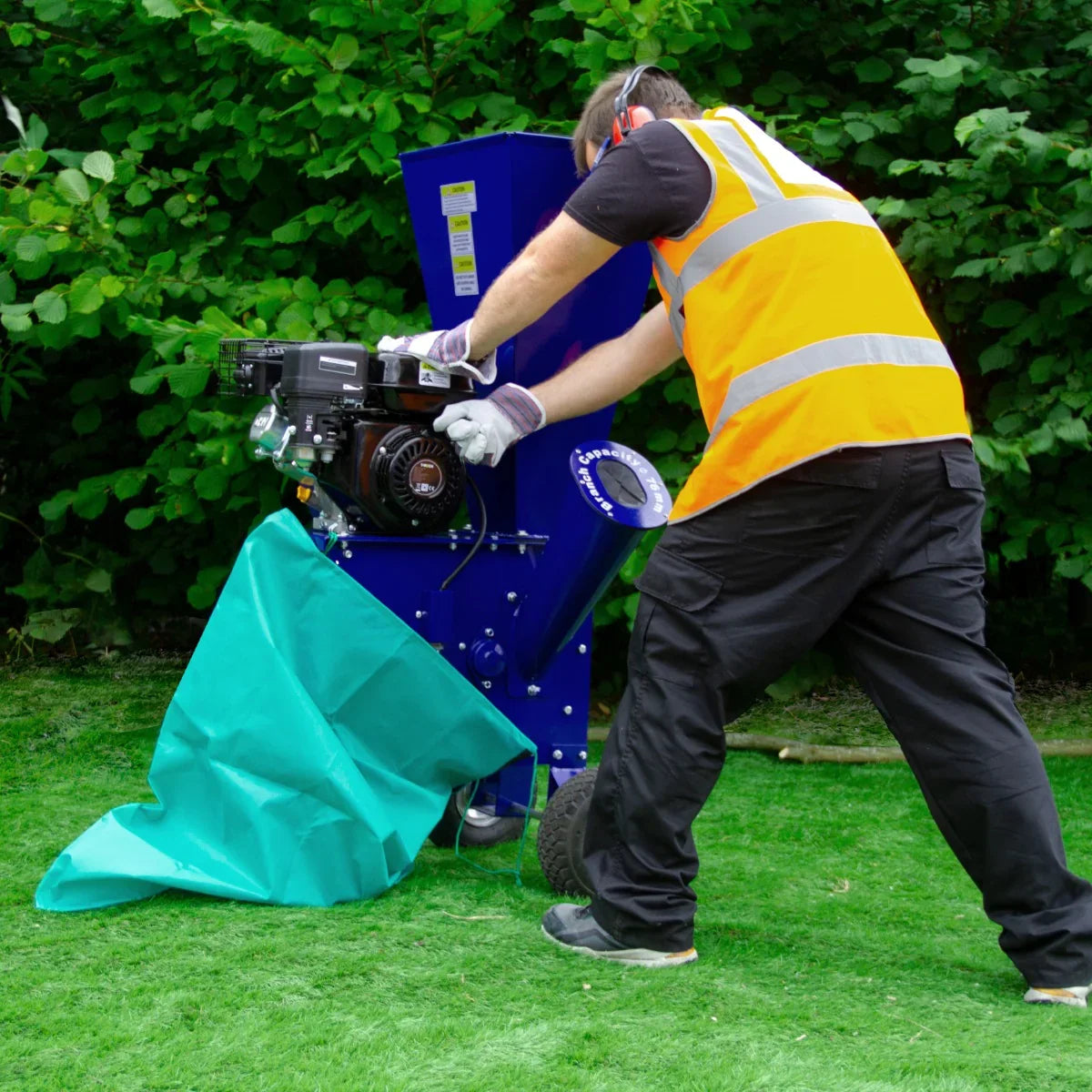 T-Mech 7HP wood chipper in action, featuring a blue body, black engine, and a worker in safety gear using a green tarp on grass