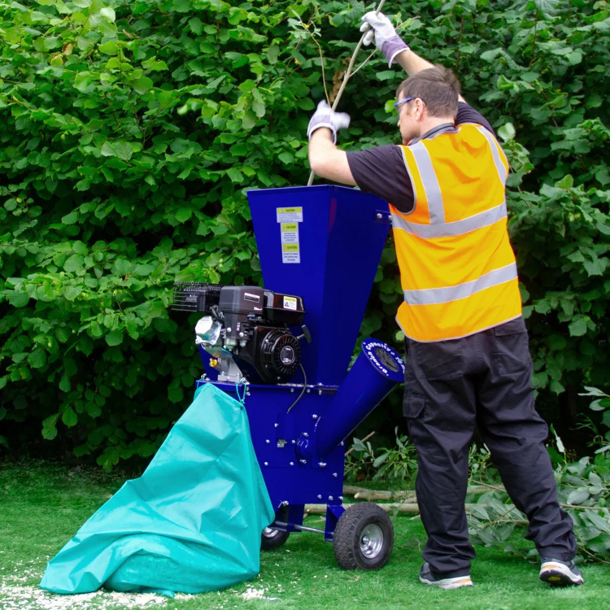 T-Mech 7HP wood chipper in blue, with a worker feeding branches into the hopper, surrounded by green foliage and grass