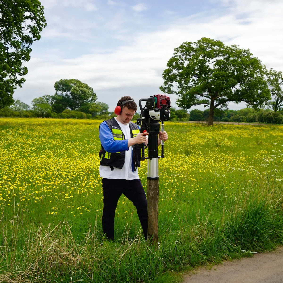 T-Mech 4 Stroke Petrol Post Driver in use by a man wearing safety gear, driving a post into a grassy field with yellow flowers