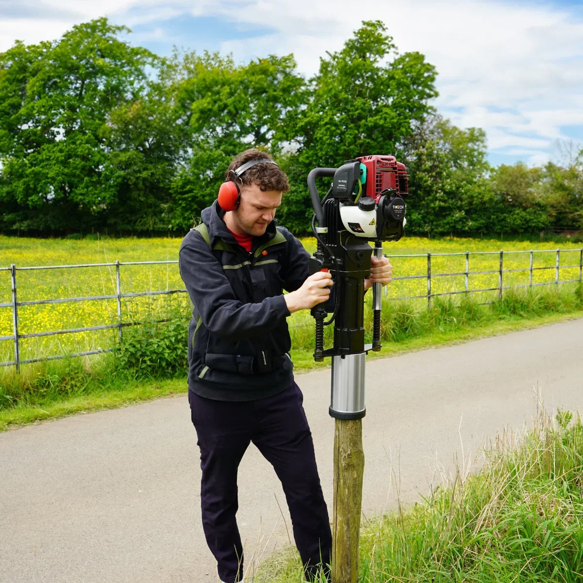 T-Mech 4 Stroke Petrol Post Driver in use, featuring a red engine, metal shaft, and a user wearing protective headphones outdoors