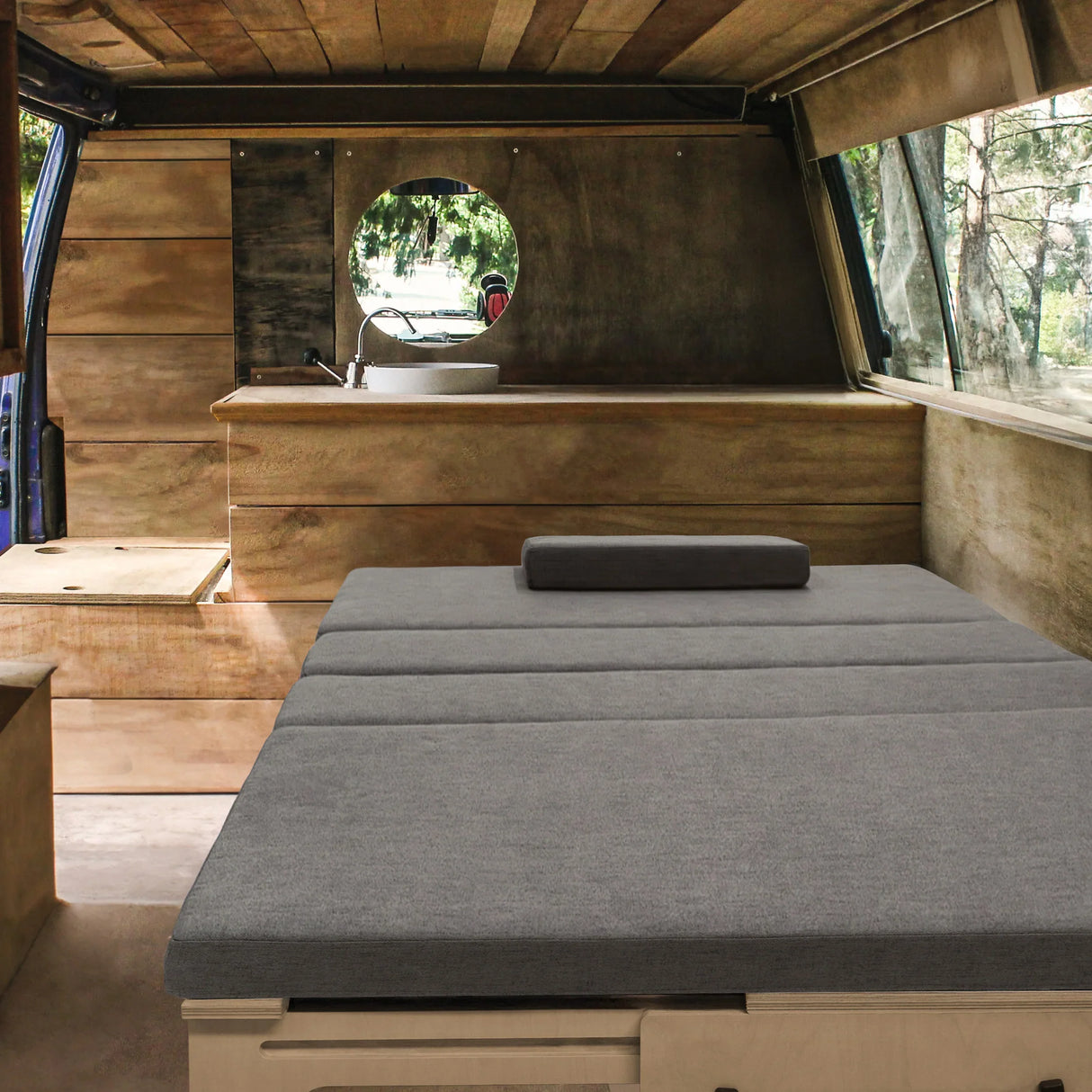 L-shaped camper van bed with a gray fabric mattress, wooden interior, and circular window, offering a cozy sleeping space