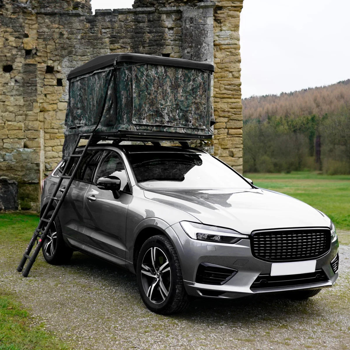 Camouflage car roof tent mounted on a silver SUV, set against a rustic stone backdrop in a grassy outdoor setting
