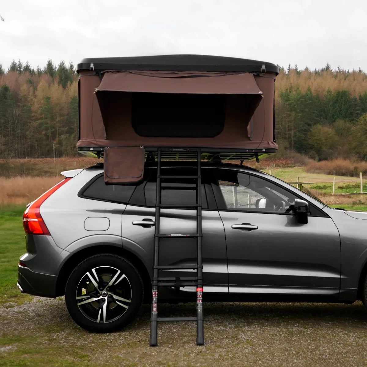 Brown car roof tent mounted on a silver SUV, featuring a sturdy ladder for access and a scenic outdoor backdrop of trees and hills