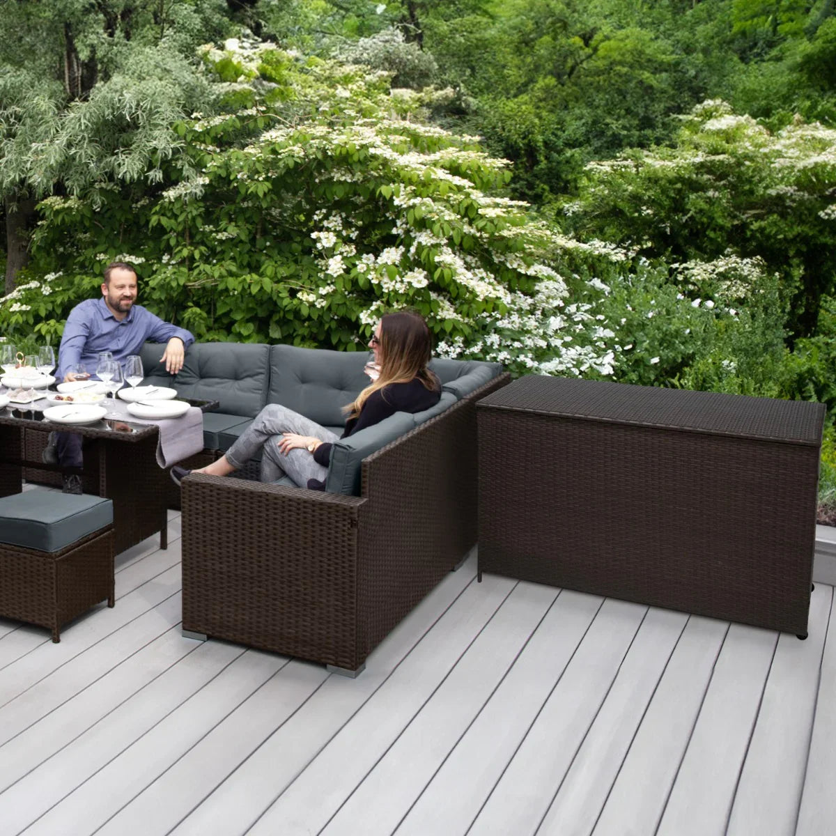 Brown rattan storage box with a textured finish, placed on a wooden deck, complementing outdoor seating and greenery in the background