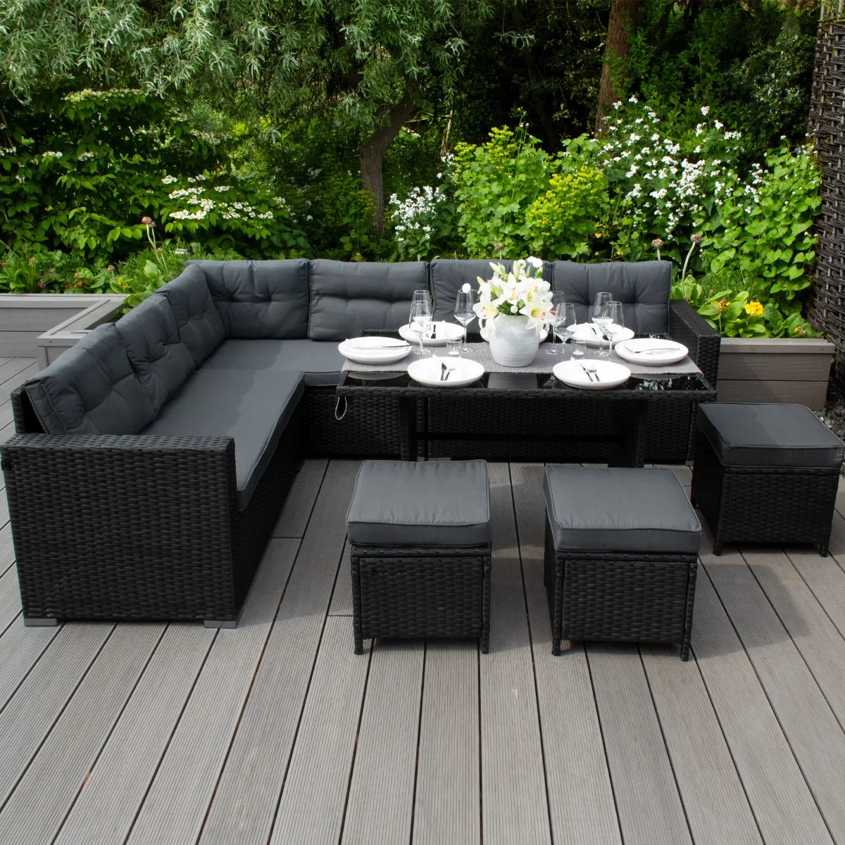 Black rattan corner set with plush cushions, matching storage box, and elegant dining setup in a lush garden setting
