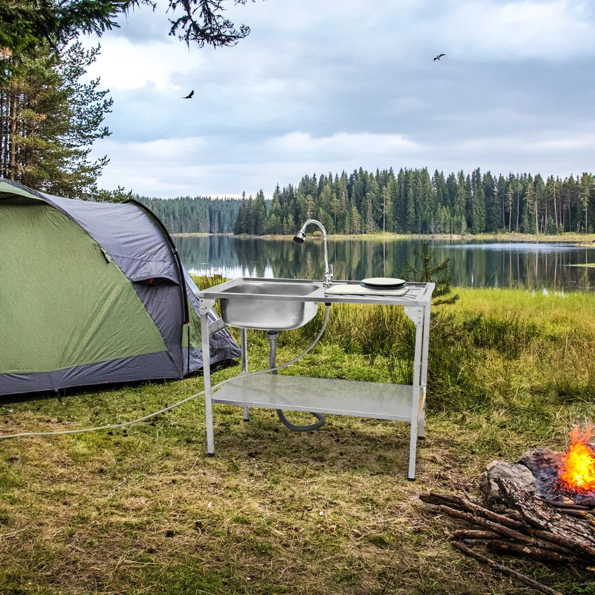 Folding camping sink with stainless steel basin, white frame, and hose, set up near a tent by a serene lake and campfire