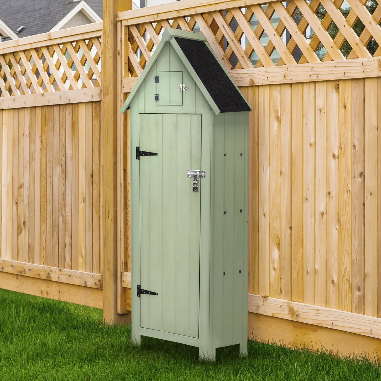 Wooden garden shed in soft green with a sloped black roof, featuring a lockable door and decorative top panel, set against a wooden fence