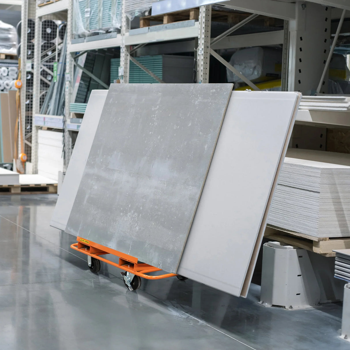 Dry wall cart with orange frame, holding large plasterboard sheets, positioned in a warehouse aisle among building materials
